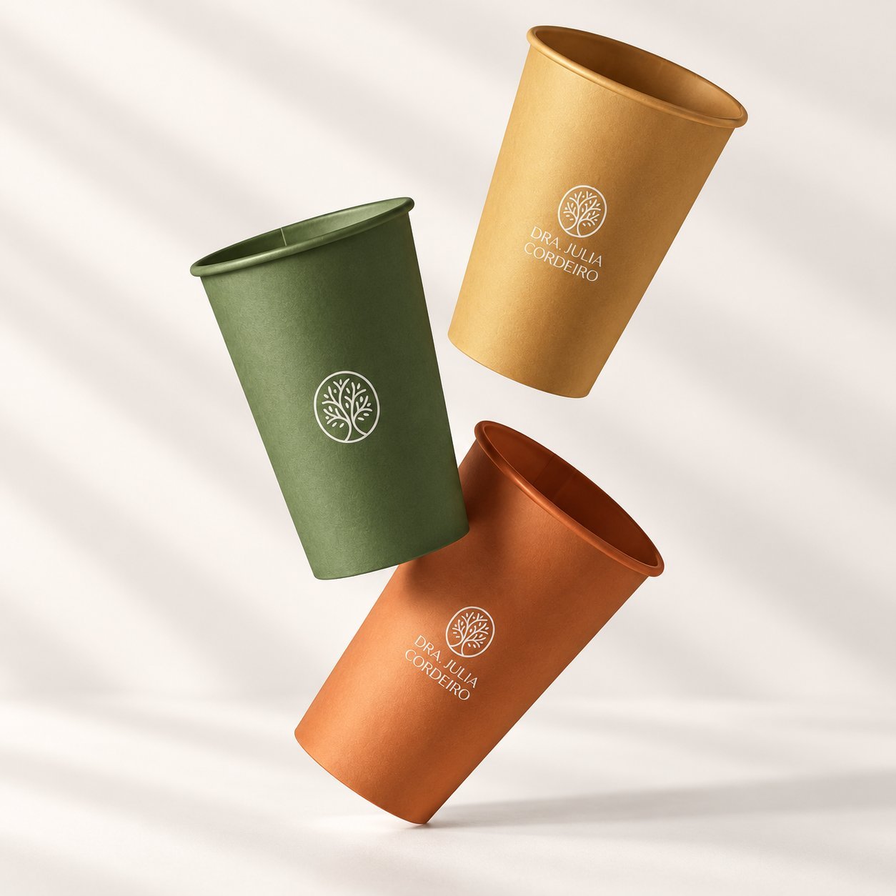 Three unprinted kraft and colored paper cups with tree logo DRA. JULIA CORDEIRO design, illustrating the pre-production sample stage where physical cups are inspected before full production.