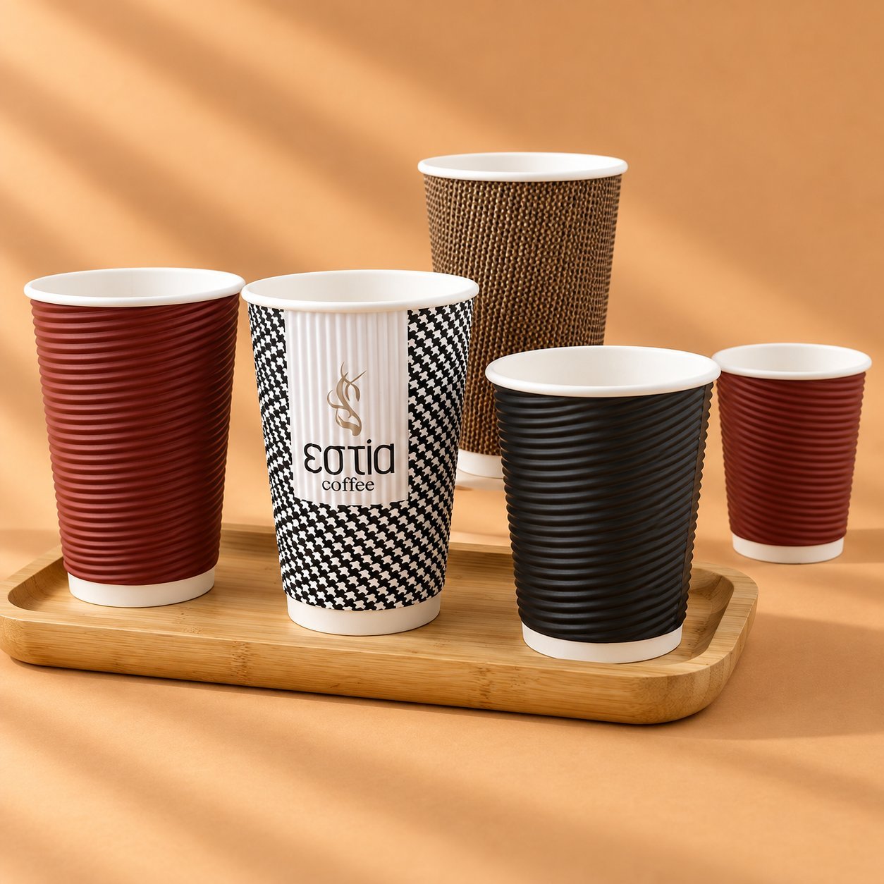 Five ripple-wall corrugated paper cups in red, houndstooth, kraft, black and red variants on a wooden tray, showing the textured surface and range of branding finishes.