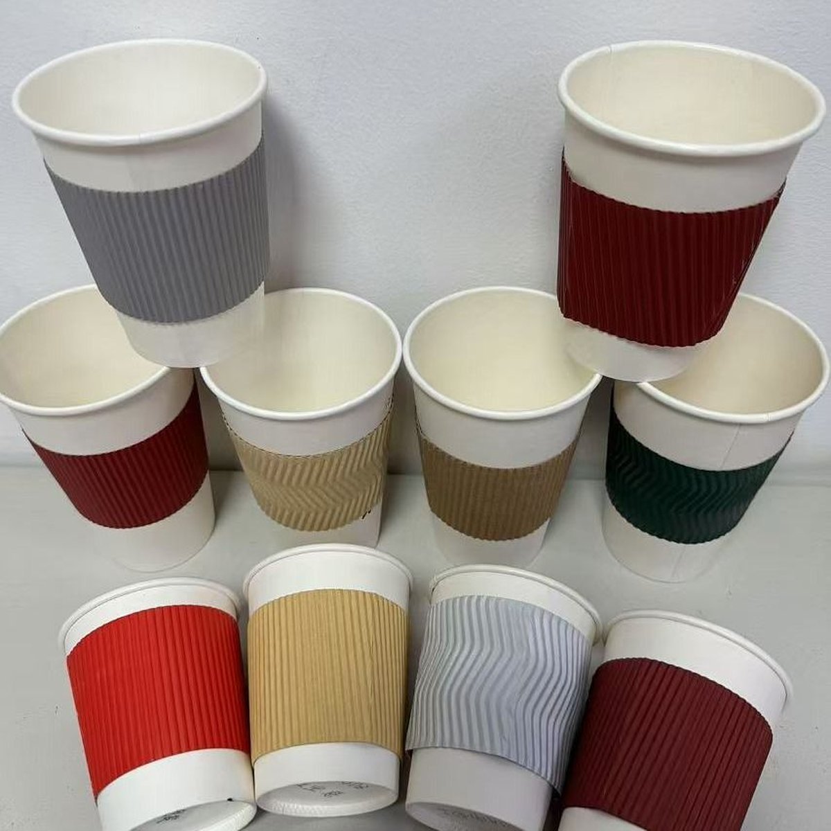 A grid of ripple-wall paper cups in several colours — burgundy, grey, tan, green, red, and white ribbed finishes — paired with their matching sleeves, illustrating how cup surface finish, ink coverage, and sleeve treatment each carry separate PFAS and heavy-metal compliance obligations under Regulation (EU) 2025/40.