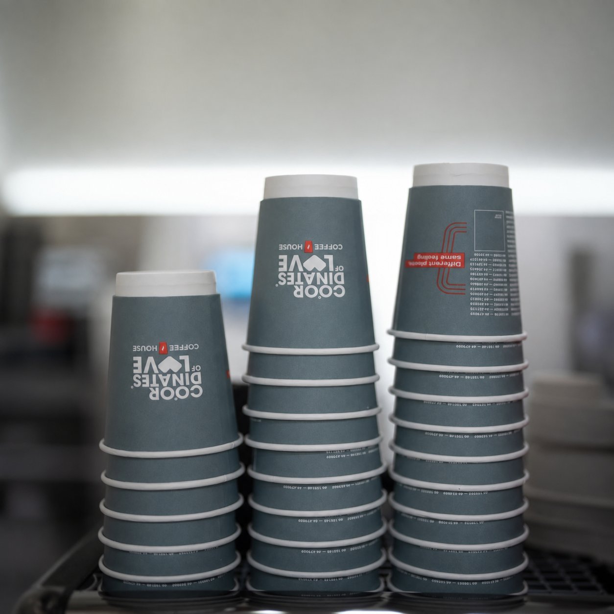 Three tall piles of branded teal paper cups printed with the 'COOR DINATES OF LOVE COFFEE HOUSE' logo and product variant text, stacked on a metal grate in a production or packing environment — a finished branded food-service SKU whose full-coverage ink and outer coating must each meet PFAS and heavy-metal thresholds under Regulation (EU) 2025/40.