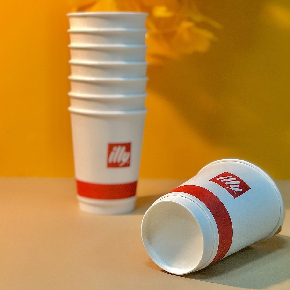 A stack of branded white paper cups with the red illy logo and a horizontal red band printed near the base, paired with a single tipped cup in the foreground — the kind of finished branded transit-ready cup format for which a Declaration of Conformity referencing Regulation (EU) 2025/40 should accompany every EU-bound shipment.