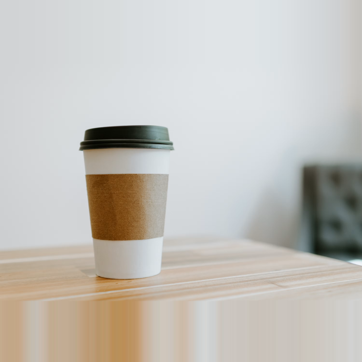 A single white paper cup with a kraft cardboard sleeve and a dark green domed lid, sitting on a light wood table with soft natural light — the kind of finished takeaway cup format that is now subject to Regulation (EU) 2025/40 substance restrictions on its coating, ink, and lid materials, applied since August 12, 2026.