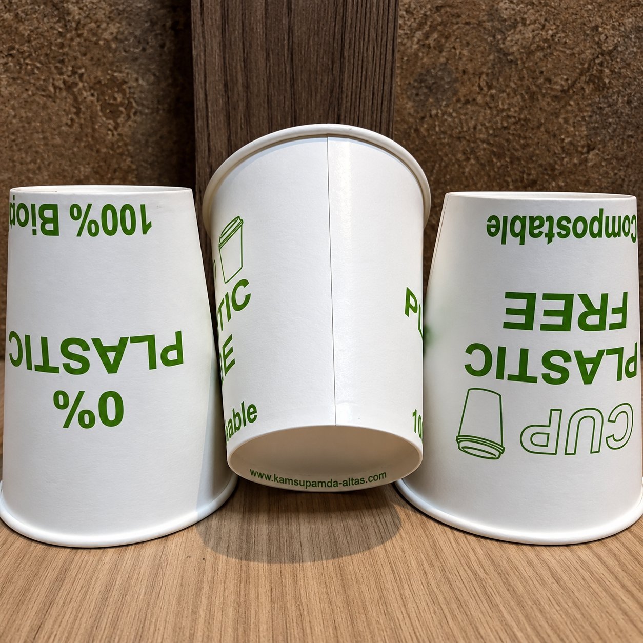 Three white paper cups arranged on a wood surface, each printed with the EU 'PLASTIC IN PRODUCT' marking in green — a clear visual reference for the marking text, box layout, and rotation options that Commission Implementing Regulation (EU) 2020/2151 specifies for partly-plastic paper cups.