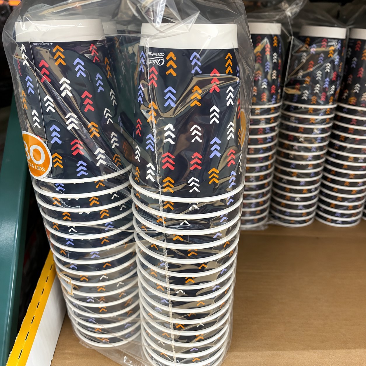 Tall stacks of dark navy paper cups with a multi-colour chevron print, shrink-wrapped in clear plastic on a warehouse shelf — a finished branded paper cup format whose plastic lining means the marking obligation applies, and where artwork and marking placement must coexist on the same external cup surface.