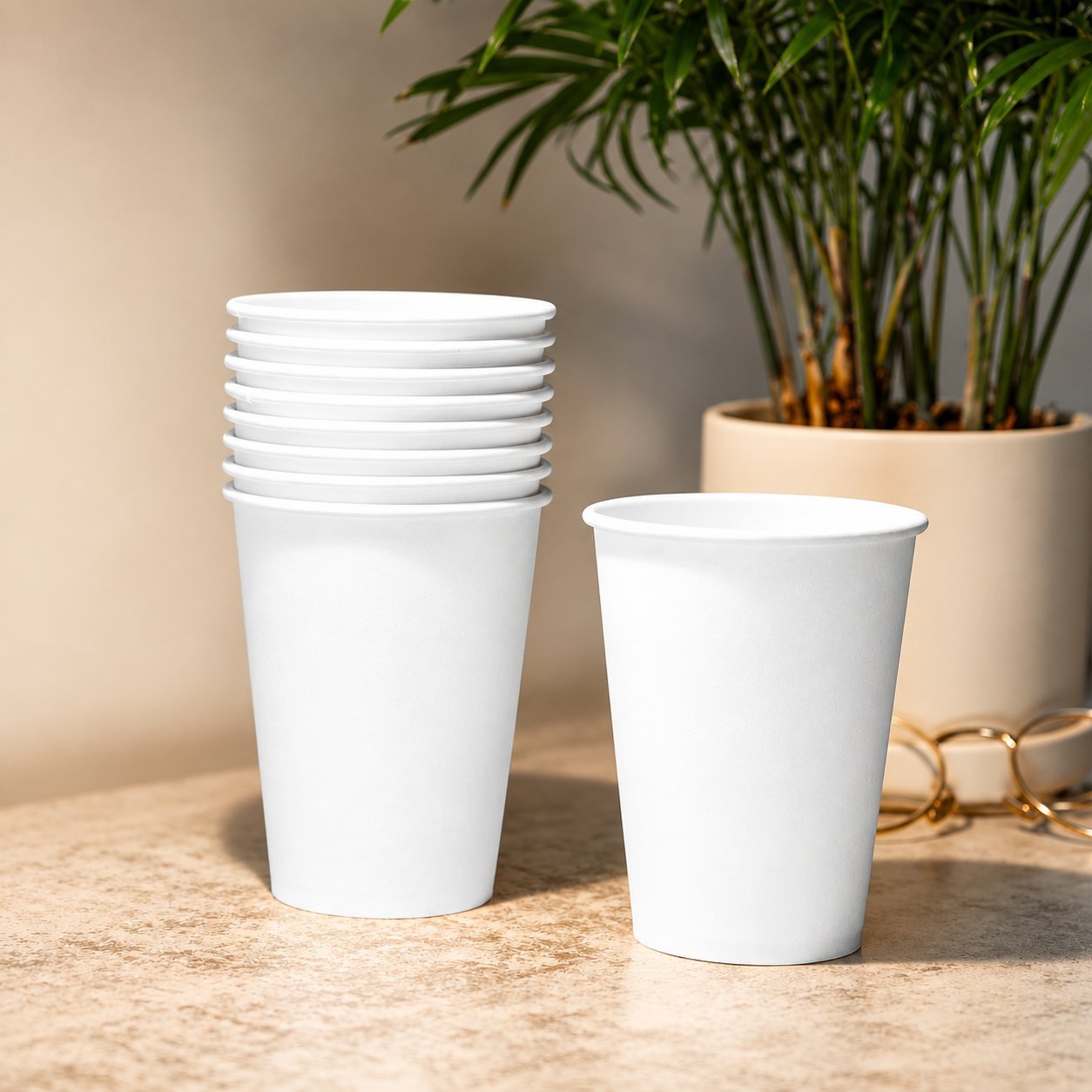 A short stack of plain white paper cups and a single tipped cup, set on a warm beige surface next to a small indoor palm in a tan pot — a calm visual reference for unprinted, minimally finished paper-cup formats that still trigger the EU marking whenever the cup carries a plastic lining or coating.